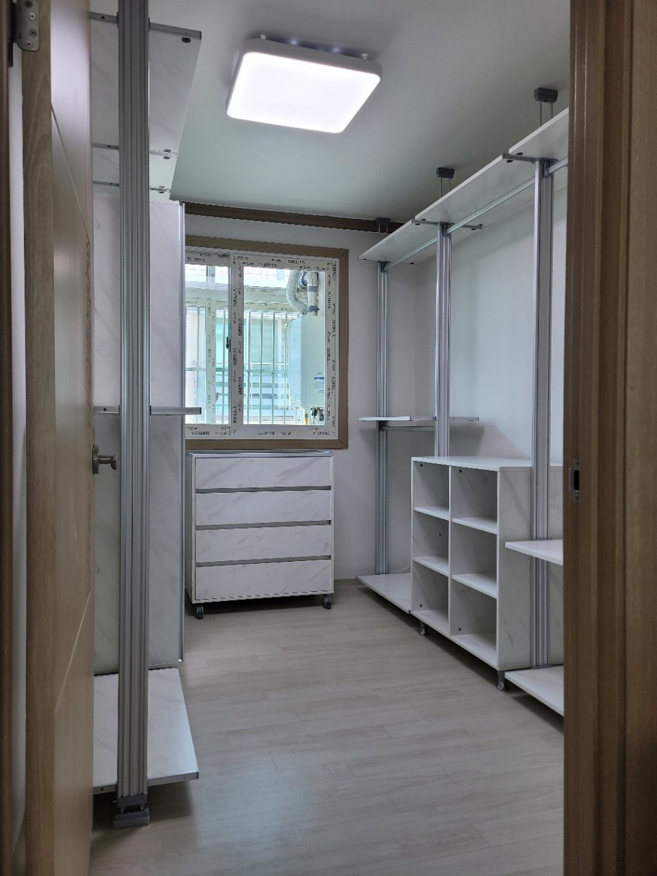 방 입구에서 본 전경. 양쪽 벽에 행거가 마주 보고 서 있고 가운데 통로 끝 창문 아래에 서랍장이 있다.